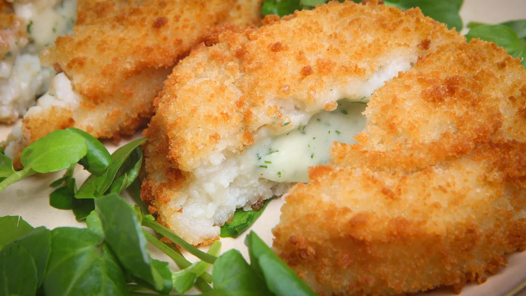 A close-up image of two golden-brown, breaded fish patties, sliced in half to reveal a creamy, green filling. The patties sit on a light-colored plate, surrounded by fresh, vibrant green pea shoots. The texture of the crispy coating contrasts with the smoothness of the filling, showcasing a appetizing dish.