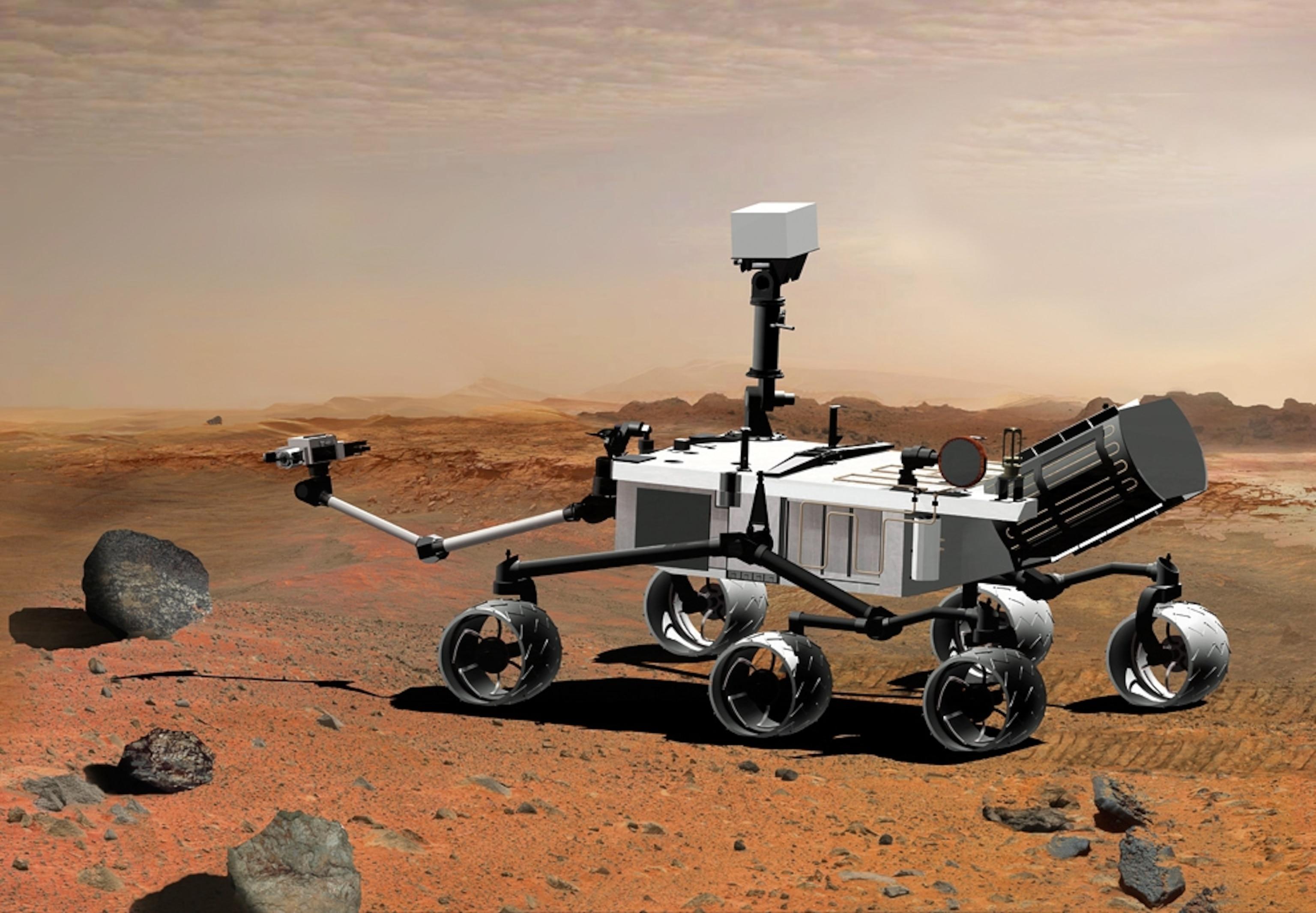 A Mars Rover is depicted on the reddish, rocky surface of Mars. The rover features six large, multi-directional wheels and a robotic arm extending toward a nearby rock. Its body is primarily white with black accents and includes various scientific instruments and a camera on top. The background shows a barren landscape under a pale sky, with additional rocks scattered throughout the scene.